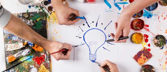 Four people draw a light bulb on a piece of paper with markers and paints.