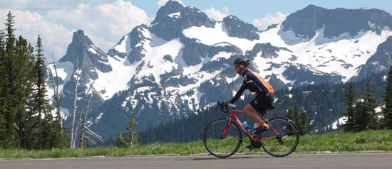 A bicyclist coasts down a mountain slope with Mount Rainier in the background.